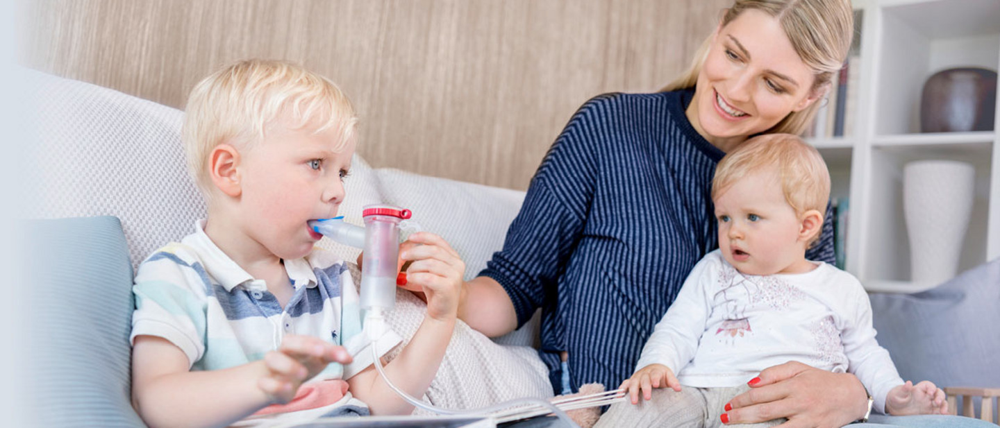 A mother helps her young daughter inhale with the PARI COMPACT2 inhaler. The girl holds the mouthpiece in her mouth. Both are sitting at a round wooden table in a bright, cheerfully furnished room with large windows.