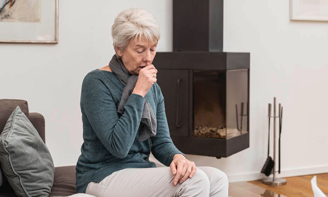 Was ist das Postnasal Drip Syndrom Ältere Frau sitzt im Wohnzimmer vor einem Kamin, trägt grauen Schal und petrolfarbenes Oberteil und hustet in ihre Hand.
