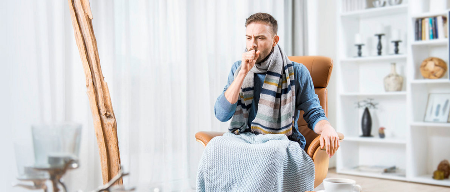 Ein junger Mann mit Schal und Decke sitzt in einem gemütlichen Wohnzimmer auf einem Sessel und hustet. Vor ihm steht ein Tisch mit einer Tasse Tee.