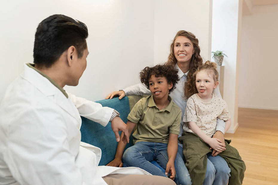A healthcare professional talks with three children and an adult seated on a couch in a clinic setting.