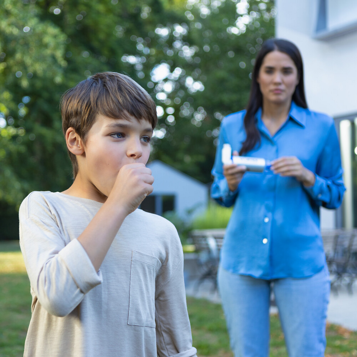 Asthma bei Kindern: Asthmaanfall Junge hustet und hält sich die Faust vor den Mund, Mutter kommt im Hintergrund herbei mir Asthmaspray und VORTEX Inhalierhilfe