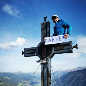 Mann sitzt auf Gipfelkreuz und hält ein Schild mit Aufschrift DANKE in der Hand