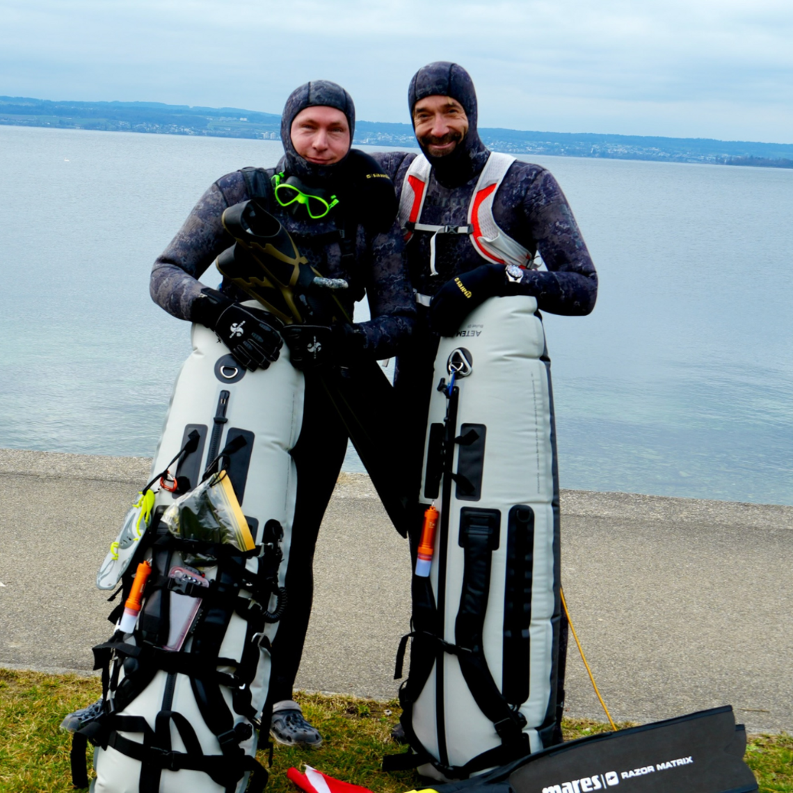 Zwei Taucher stehen nebeneinander am Ufer mit Ausrüstung und Rucksack