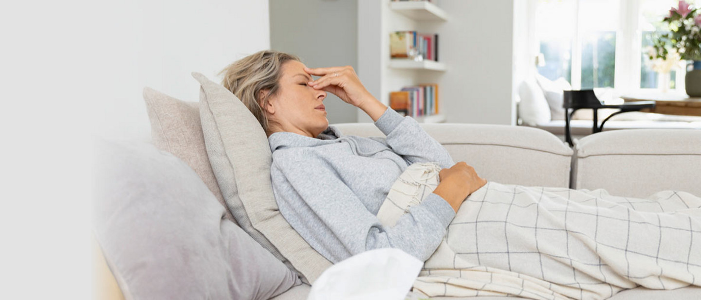Eine Frau liegt krank auf einem Sofa in einem Wohnzimmer. Sie ist mit einer Decke zugedeckt. Sie hält sich die Stirn und wirkt erschöpft. Vor ihr liegt eine Packung Taschentücher.