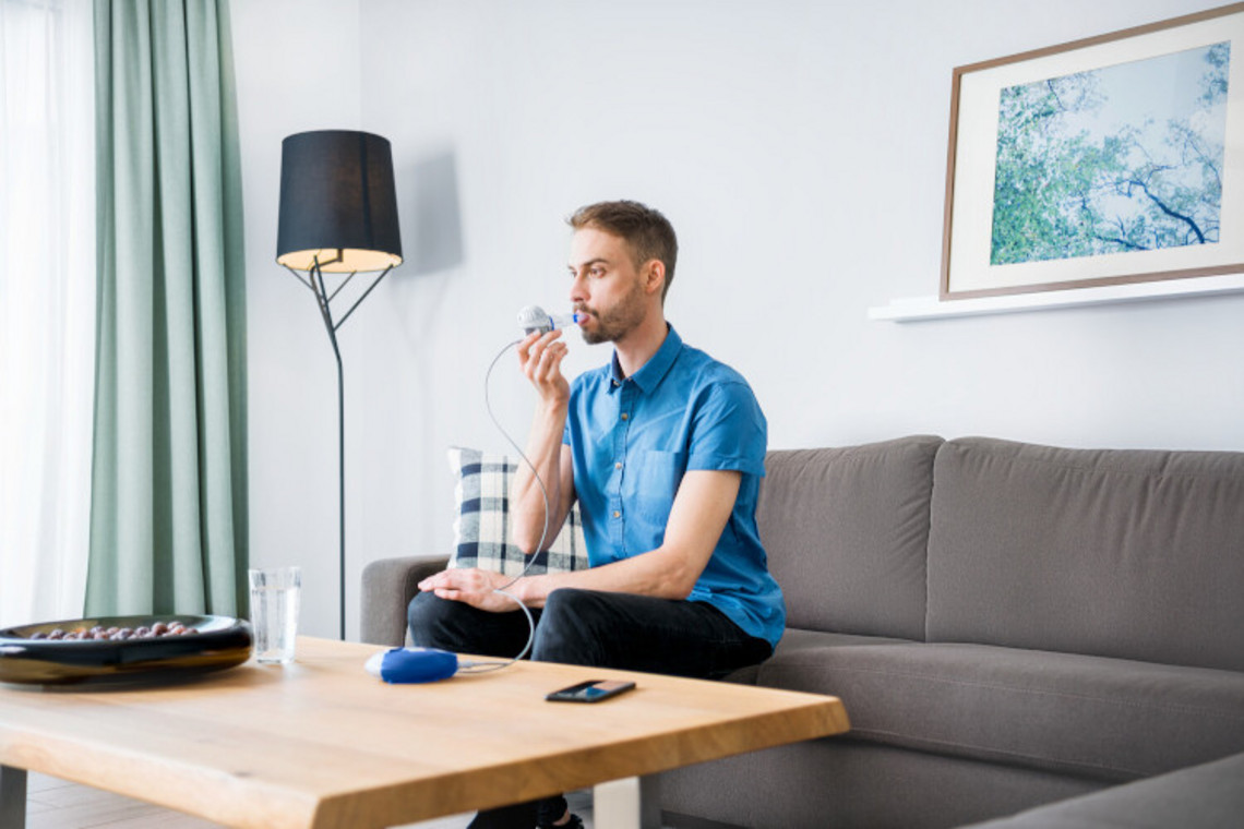 Zeit sparen bei der Atemtherapie: eFlow®rapid Mann inhaliert mit Inhalationssystem auf dem Sofa sitzend