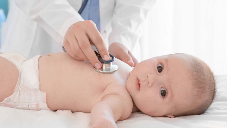 Säugling liegt auf Liege und schaut in die Kamera, Arzt hält Stethoskop auf seine Brust