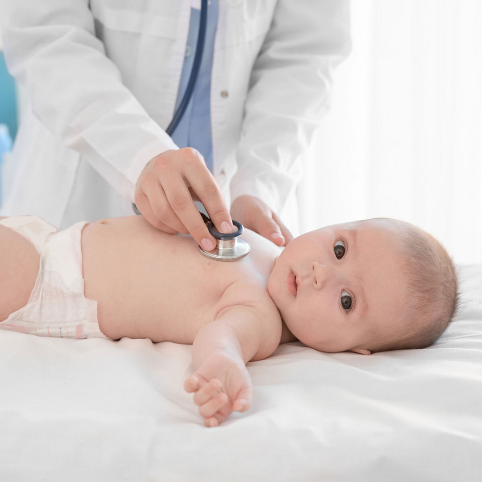 Säugling liegt auf Liege und schaut in die Kamera, Arzt hält Stethoskop auf seine Brust