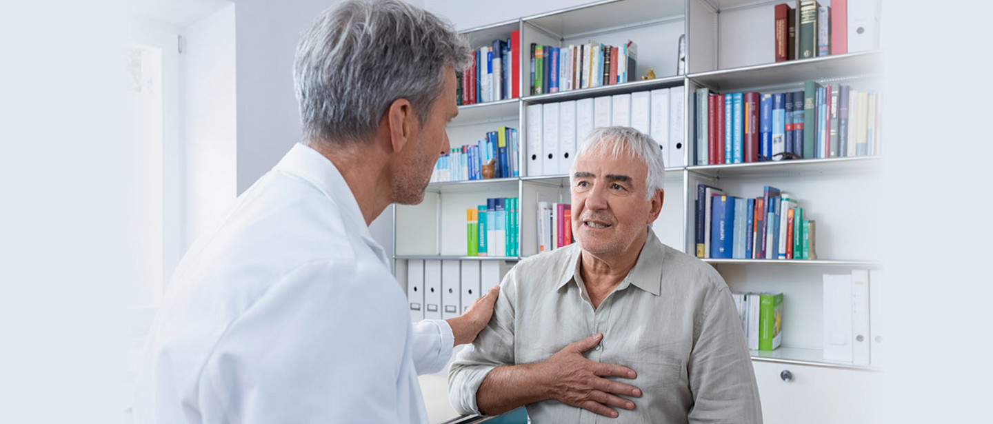 An elderly man sits in a doctor's office, holds his hand on his chest and talks to a doctor in a white coat who places a reassuring hand on his shoulder.