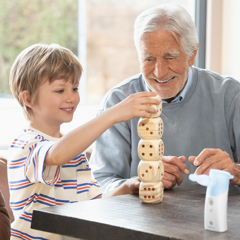 A child is stacking wooden blocks at a table; another person is sitting next to him; the PARI BOY Go inhaler is in the foreground.