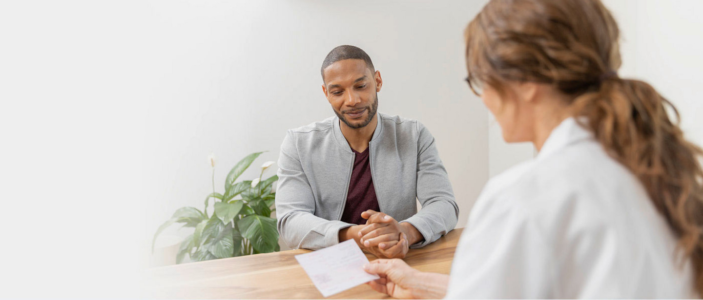 Ein Mann sitzt entspannt an einem Holztisch und blickt auf ein Rezept, das ihm eine Ärztin in weißem Kittel übergibt. Im Hintergrund steht eine grüne Zimmerpflanze, die Umgebung wirkt freundlich und modern.