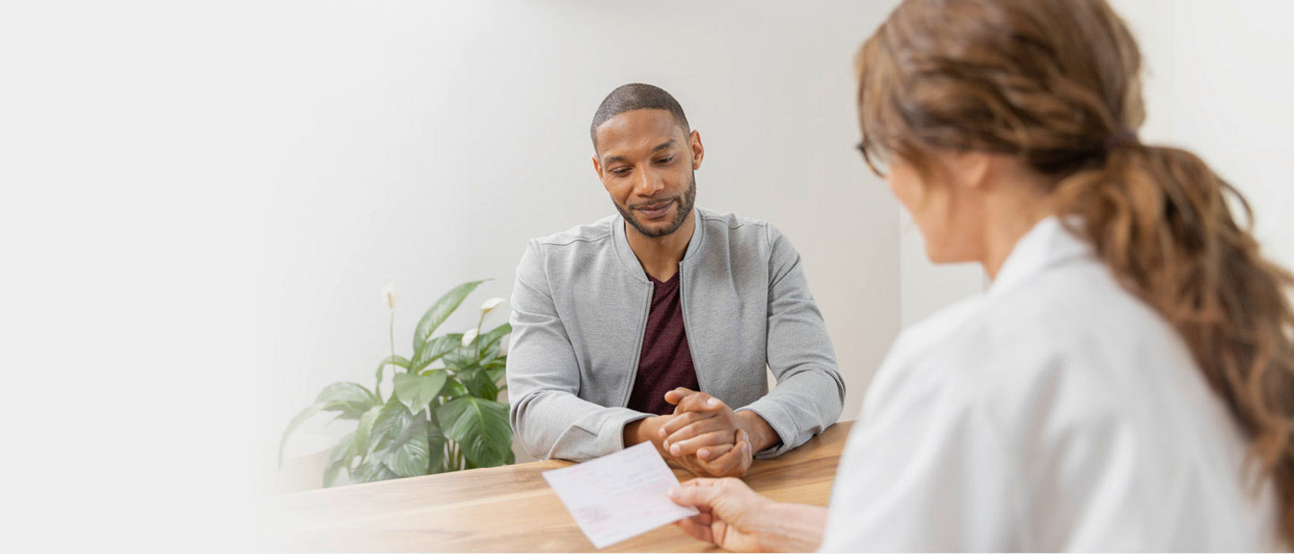 Ein Mann sitzt an einem Holztisch und betrachtet ein Rezept, das ihm eine Ärztin im weißen Kittel überreicht.