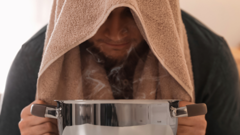 Junger Mann sitzt vor Topf mit dampfendem Wasser und Handtuch über dem Kopf, um zu inhalieren