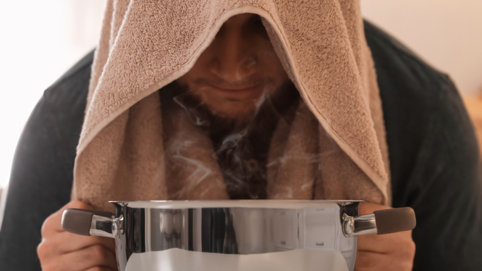 Junger Mann sitzt vor Topf mit dampfendem Wasser und Handtuch über dem Kopf, um zu inhalieren