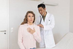 Junge Frau sitzt beim Arzt, der sie am Rücken mit Stethoskop untersucht