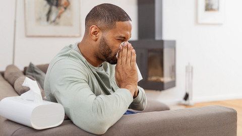 Junger Mann sitzt auf Couch und putzt sich die Nase
