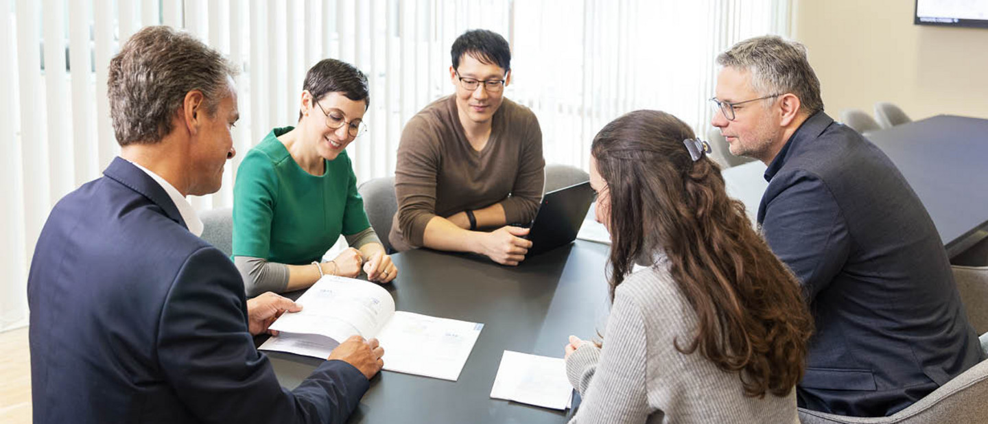 Besprechung mit sechs Personen an einem Konferenztisch in modernem Büro, Fokus auf gemeinsames Dokument und Austausch.