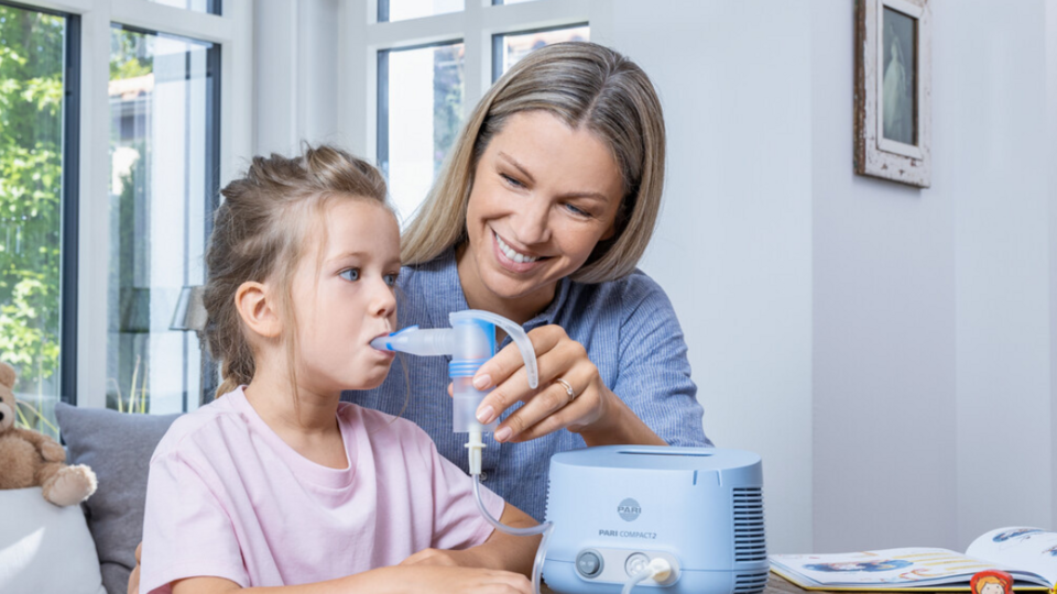 Home remedies against a cough: Loosen mucus naturally, ease a dry cough Junge Mutter sitzt mit Tocher am Tisch und hilft ihr beim Inhalieren mit einem PARI COMPACT2 Inhalationsgerät