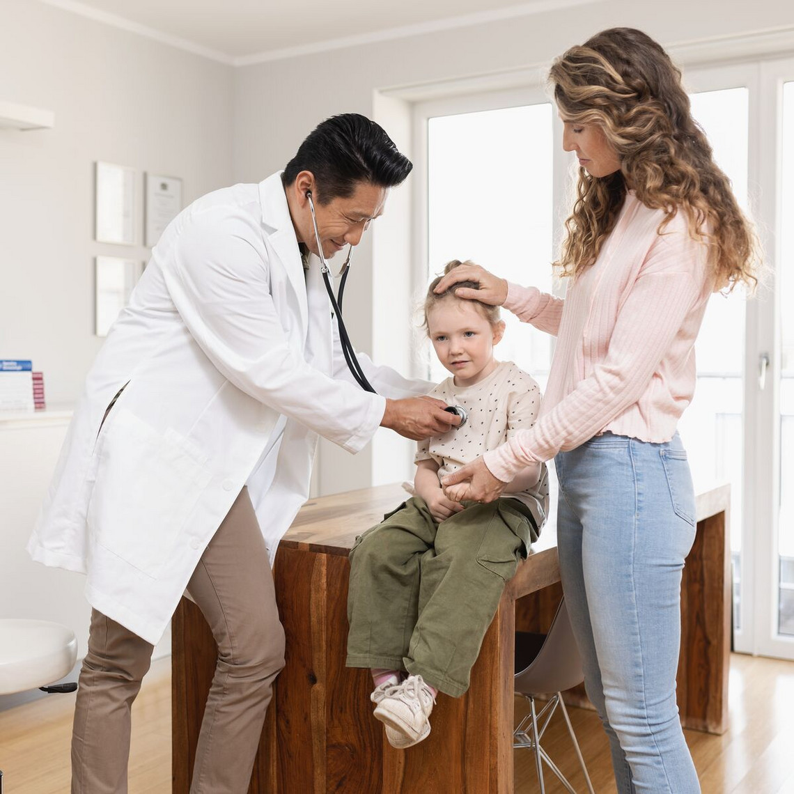 Girl is examined by doctor with stehoscope, mother stands next to her and holds her hand reassuringly on the girl's head