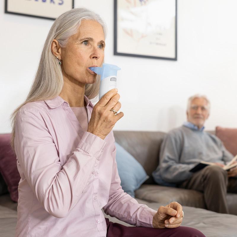 A person is sitting in the living room using a PARI BOY Go inhaler with a mouthpiece; another person is in the background.