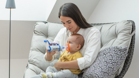 Young mother sits in an armchair with her baby on her lap and helps him inhale asthma spray using the VORTEX spacer