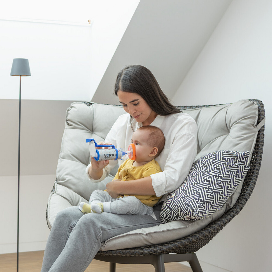 Young mother sits in an armchair with baby on her lap and helps him to inhale with asthma spray and a spacer