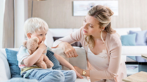 Kleiner Junge sitzt mit Kuscheltier auf der Couch, Mutter sitzt besorgt davor