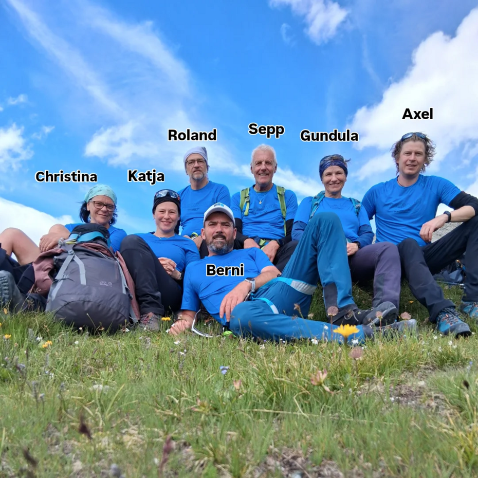 Eine Gruppe von sieben Menschen mit blauen T-Shirts sitzt auf einer Wiese und lächelt in die Kamera