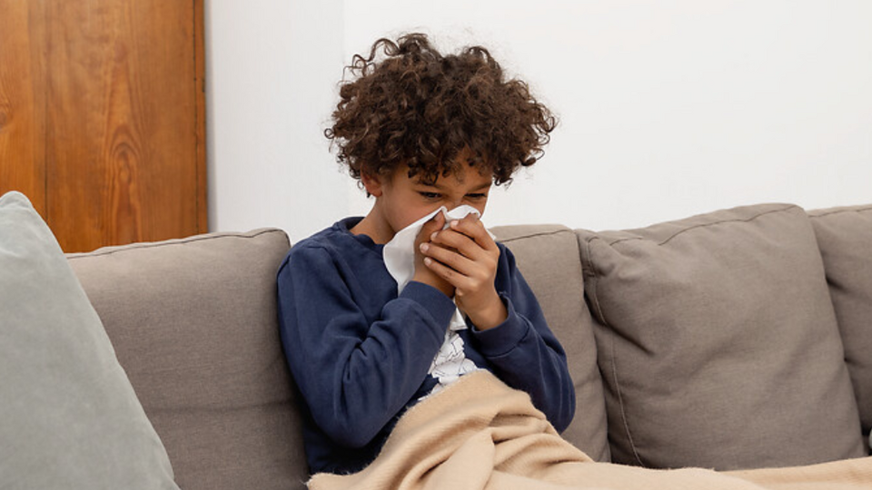 Junge sitzt mit Decke auf der Couch und schnäuzt in ein Taschentuch