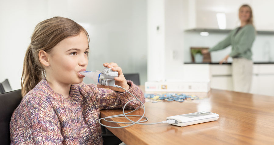 A girl sits on a chair in front of a table and inhales with the eFlow®rapid.