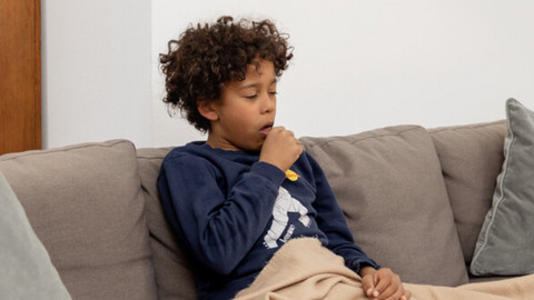 Junge sitzt auf Couch mit Decke und hustet in die Faust