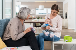 Mother helps baby inhale with PARI BOY Junior inhalation device while grandmother sits beside and watches