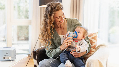 Junge Mutter hat Baby auf dem Schoß, Baby inhaliert mit Vernebler und Maske