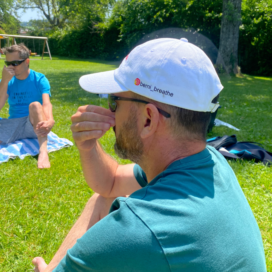 Mann sitzt mit einer Cap auf der Wiese und führt die Wechselatmung durch
