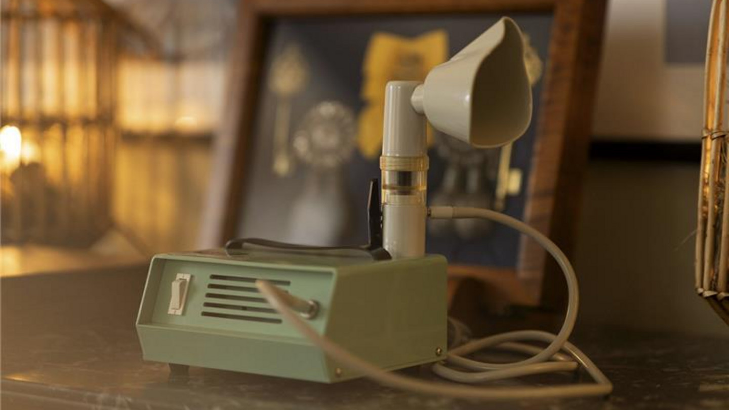 Vintage-Inhalationsgerät mit Mundstück und Schlauch steht auf einem Tisch vor einem gerahmten Bild.