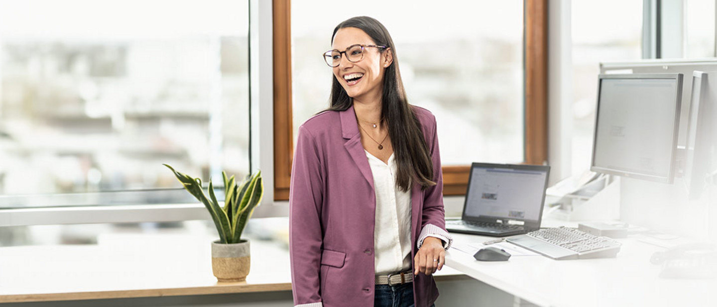 A smiling woman wearing glasses and a purple blazer stands in a modern office next to a desk with several monitors and a laptop.