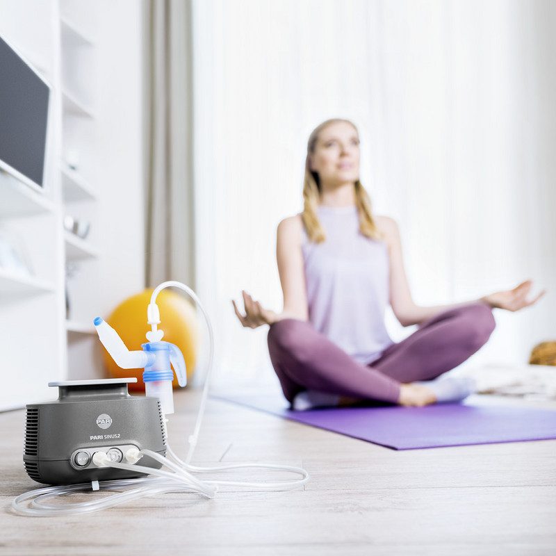 Eine junge Frau sitzt im Wohnzimmer im Schneidersitz auf einer Jogamatte und meditiert. Im Vordergrund steht ein PARI SINUS2 Inhalationsgerät.
