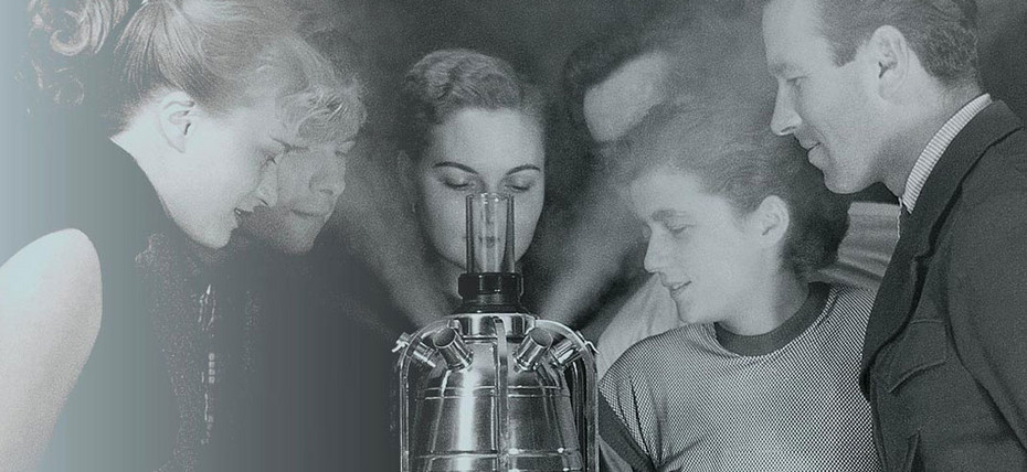 Historisches Schwarzweißfoto: Mehrere Personen im Hintergrund, im Vordergrund ein Glasgerät, aus dem feiner Nebel austritt. Historisches Schwarzweißfoto: Mehrere Personen im Hintergrund, im Vordergrund ein Glasgerät, aus dem feiner Nebel austritt.
