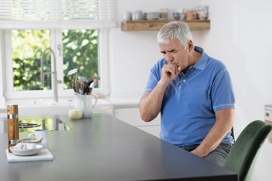 FAQ Inhalation bei COPD: Mann hustet Älterer Mann in gestreiftem Hemd und Schal sitzt im Wohnzimmer, hält sich beim starken Husten eine Hand vor den Mund und die andere an die Brust.