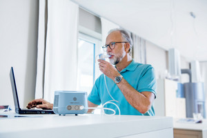 Älterer Mann inhaliert mit Inhalationssystem, während er am Tisch am Laptop arbeitet