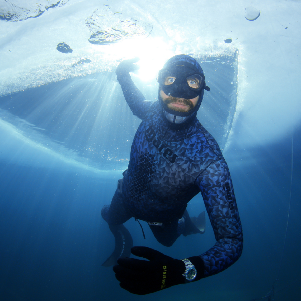Taucher von unten aus der Tiefe fotografiert, über ihm eine Eisschicht über der Wasseroberfläche