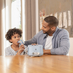 Mythos Dampfinhalation: PARI COMPACT2 Junge sitzt am Tisch und inhaliert mit dem PARI COMPACT2 Inhalationsgerät, Vater sitzt lächelnd daneben