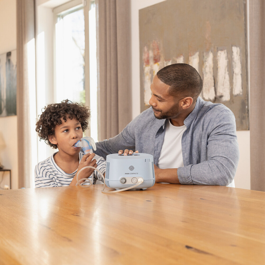 Junge sitzt am Tisch und inhaliert mit dem PARI COMPACT2 Inhalationsgerät, Vater sitzt lächelnd daneben