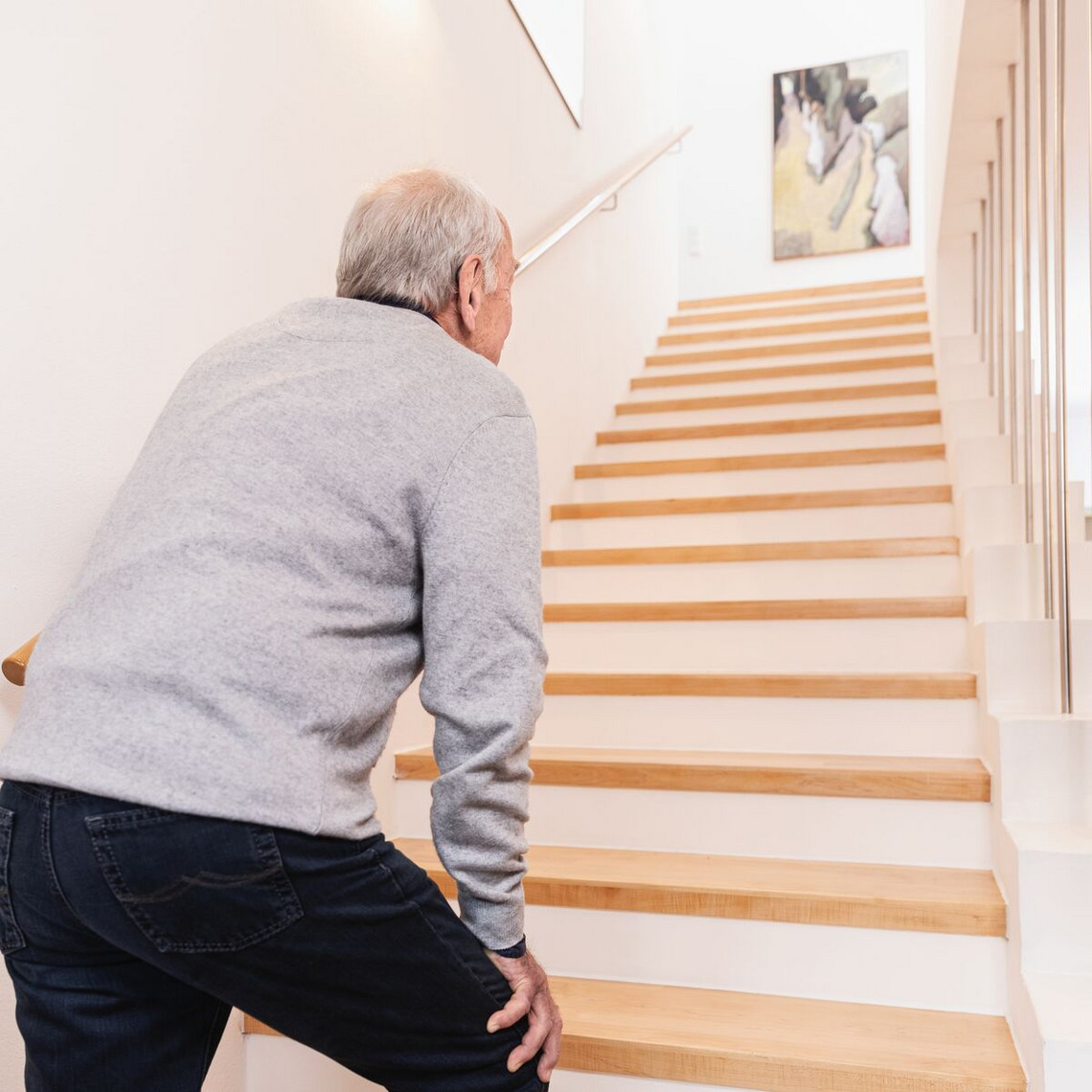 Photograph of an elderly man from behind, standing at the foot of a staircase and looking upwards with his hand resting on his thigh