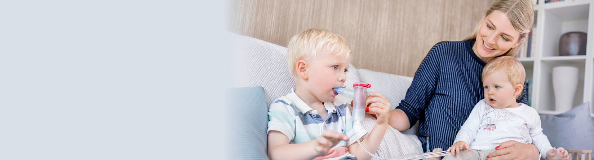 A mother helps her young daughter inhale with the PARI COMPACT2 inhaler. The girl holds the mouthpiece in her mouth. Both are sitting at a round wooden table in a bright, cheerfully furnished room with large windows.
