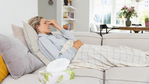Junge Frau liegt auf Couch und hält sich die Stirn