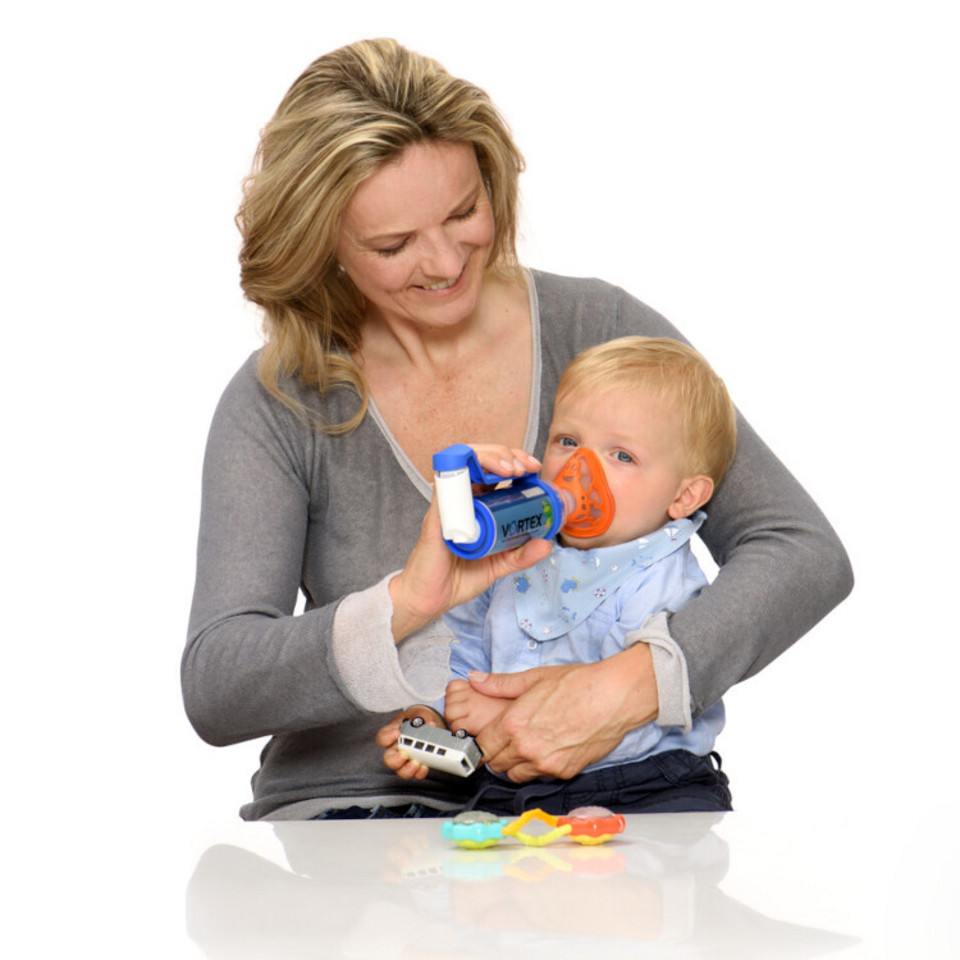 Mother holding asthma spray with spacer and mask in front of baby’s face on her arm