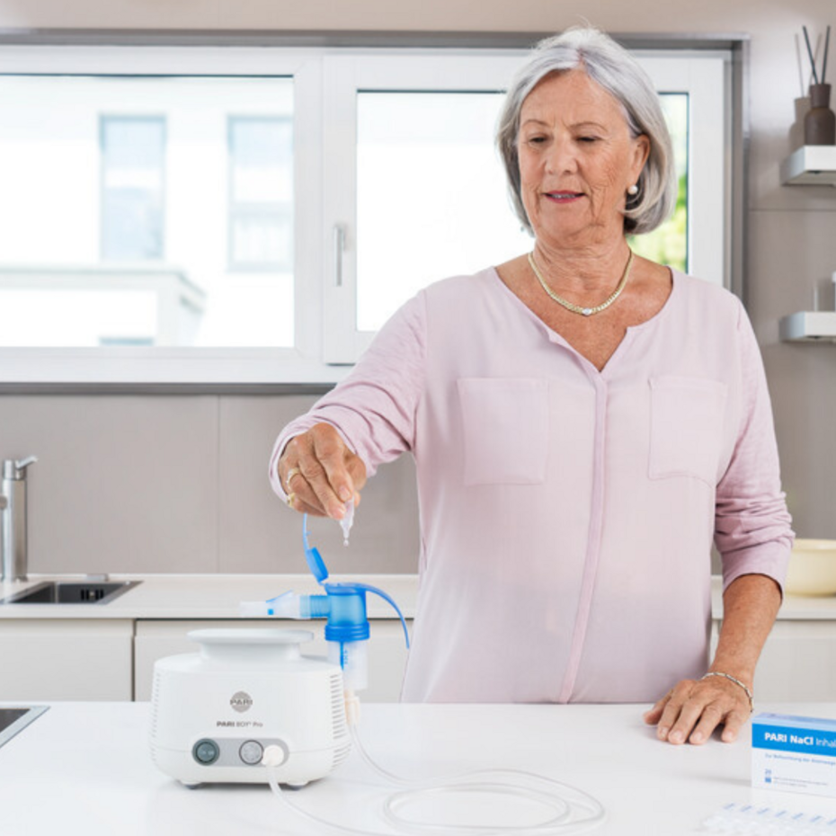 Ältere Frau steht in der Küche am Tisch und füllt Inhalationslösung in das PARI BOY Pro Inhalationsgerät
