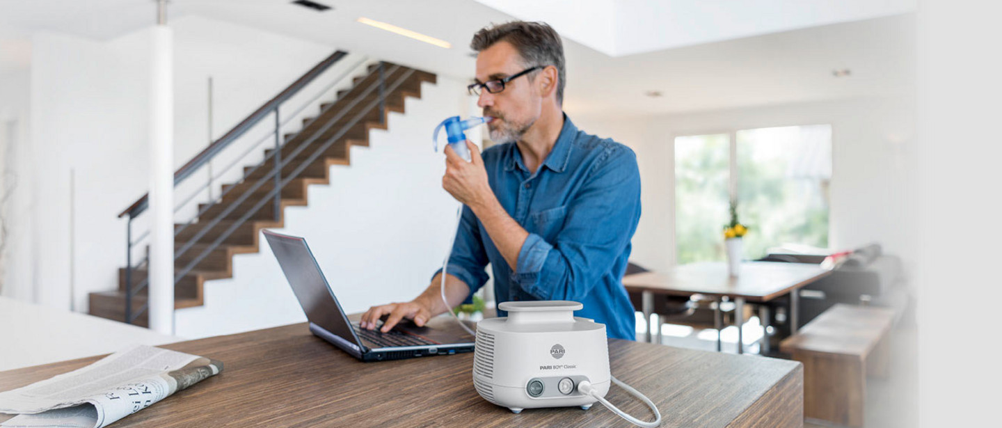 Ein Mann mittleren Alters mit Brille und grauen Haaren sitzt an einem modernen Tisch in einem hellen, offenen Wohnraum. Er trägt ein blaues Hemd, arbeitet am Laptop und inhaliert mit dem PARI LC SPRINT. Daneben steht ein PARI BOY Classic mit Schlauch, eine Zeitung liegt auf dem Tisch. Im Hintergrund sind eine Treppe, eine Glasbalustrade und eine Sitzecke mit großen Fenstern zu sehen.