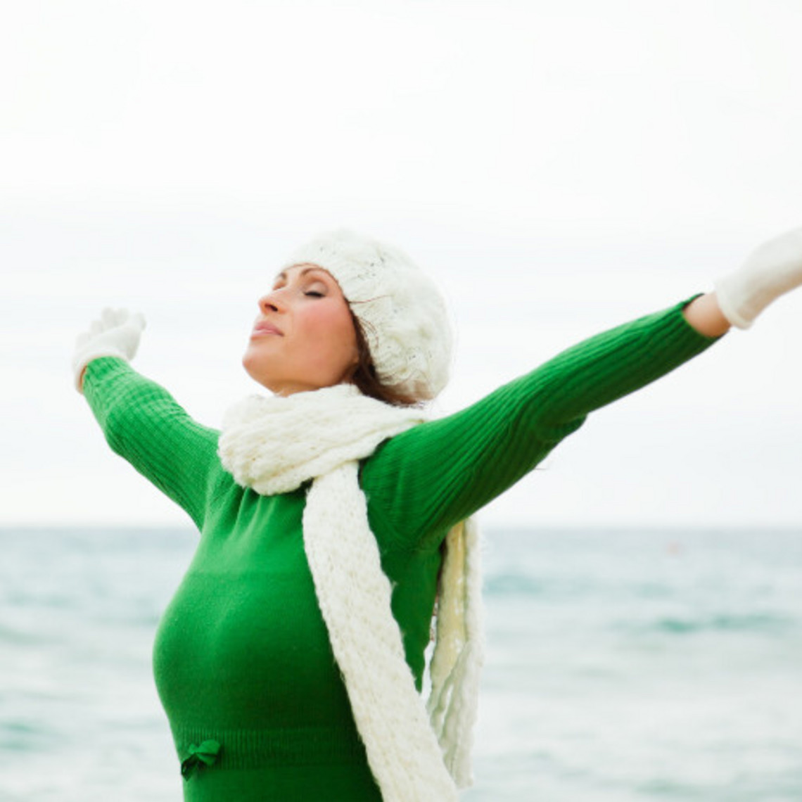 Junge Frau mit Mütze und Schal und ausgestreckten Armen atmet tief durch, im Hintergrund das Meer