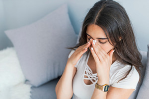 Junge Frau sitzt auf dem Sofa und hält beide Hände an die Nasenwurzel